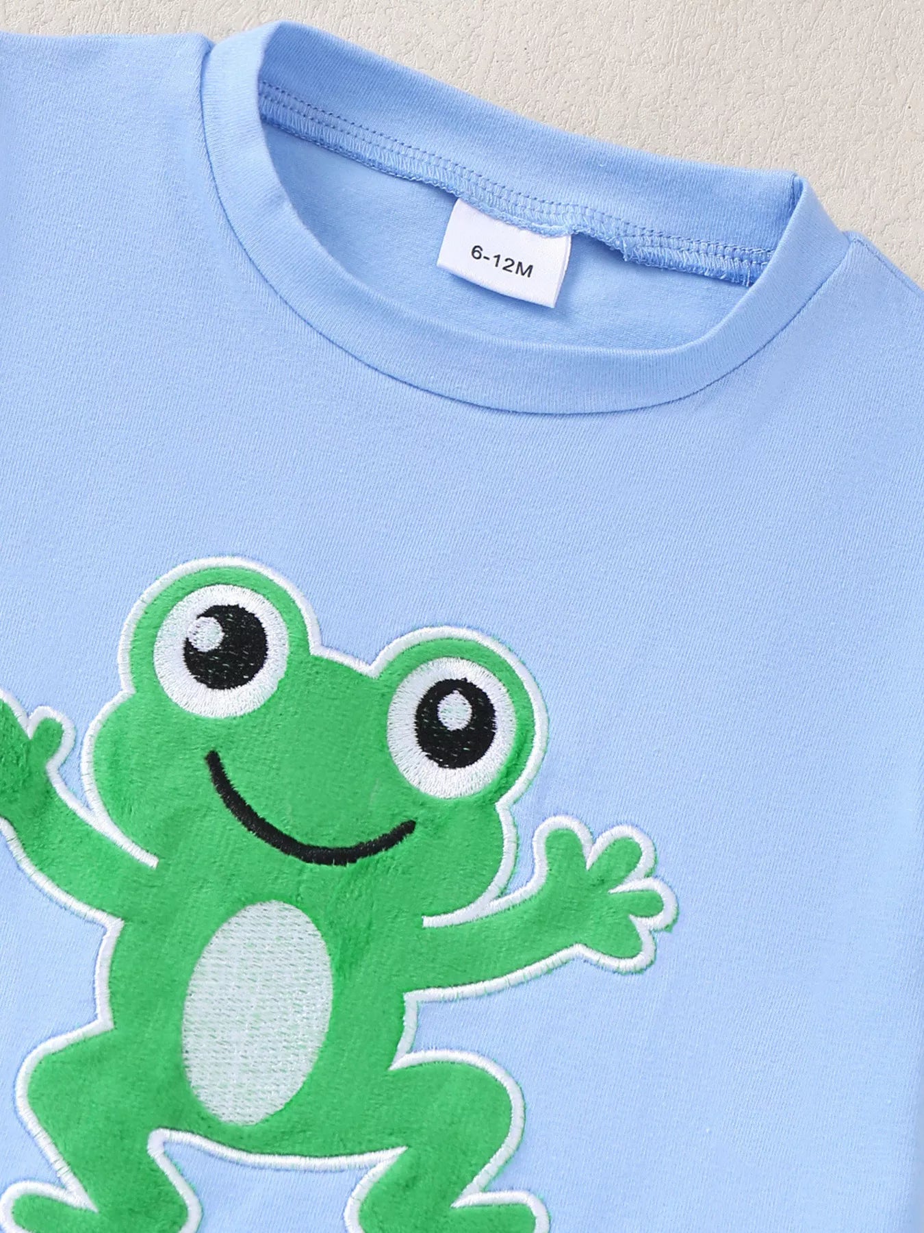 Blue baby T-shirt with green frog appliqué and round neckline, upper front close-up
