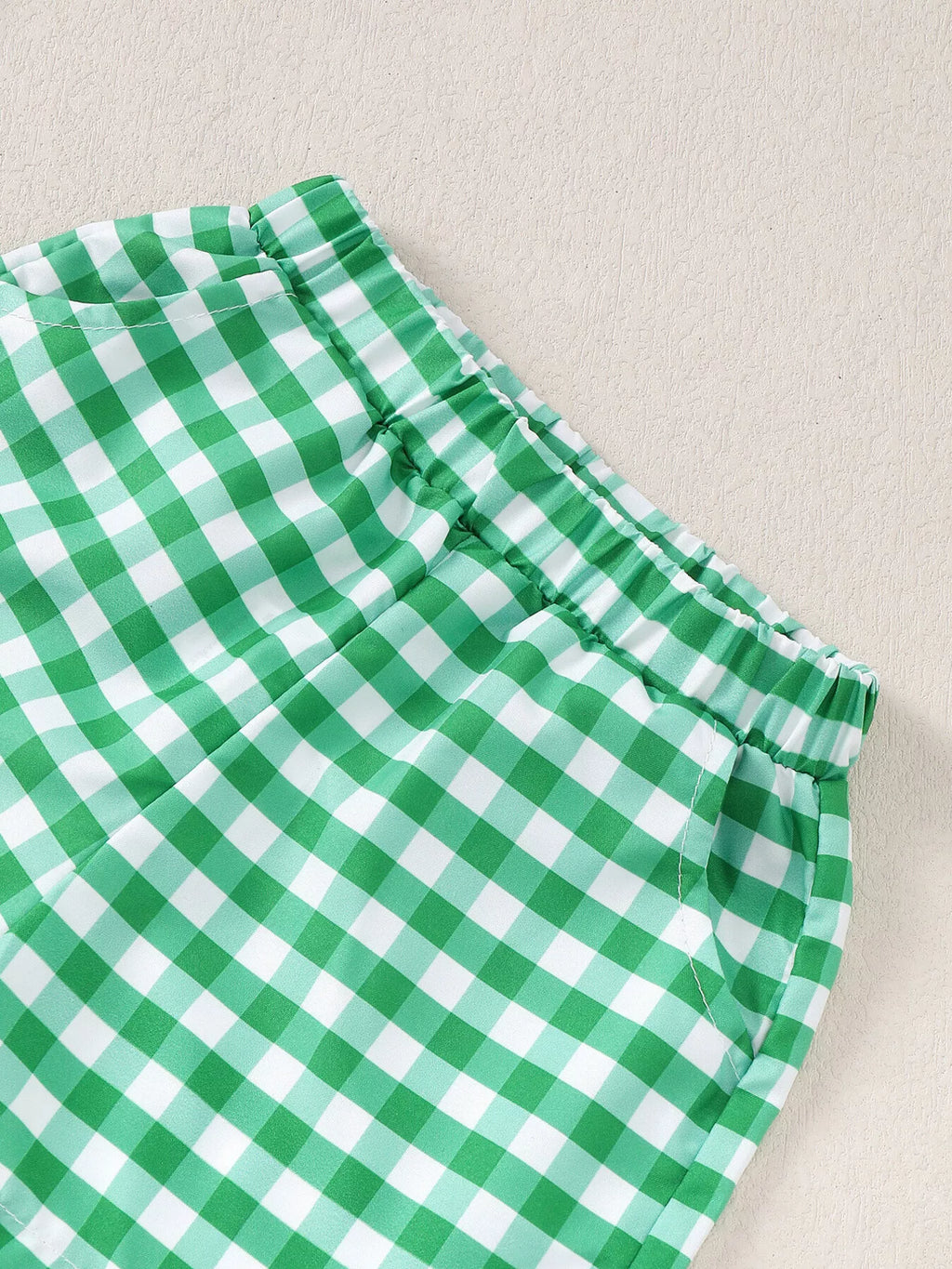 Green gingham baby shorts with elastic waistband and curved side pockets, close-up view