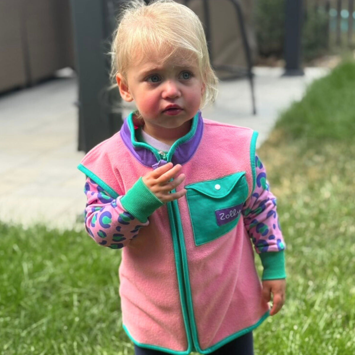 Bubblegum pink toddler fleece vest with purple collar and teal chest pocket, modeled outdoor front view