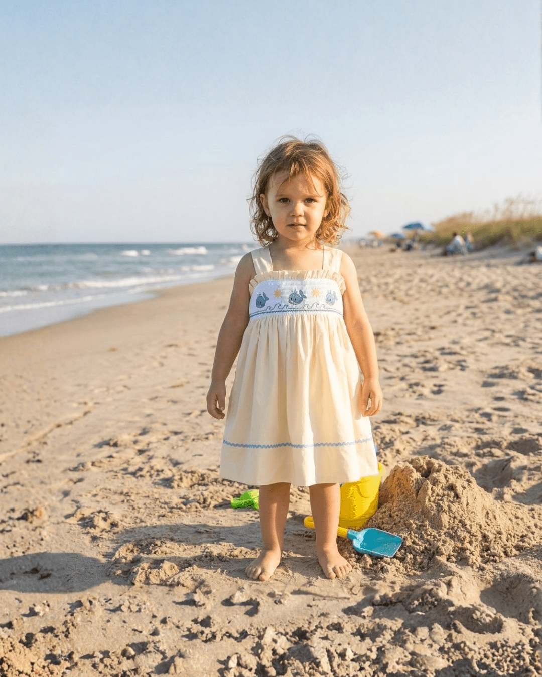 Buttercream yellow kids whale smocked dress with criss-cross straps, modeled beach view