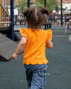 Orange kids flutter-sleeve top with white trim and scalloped hem, rear playground lifestyle view