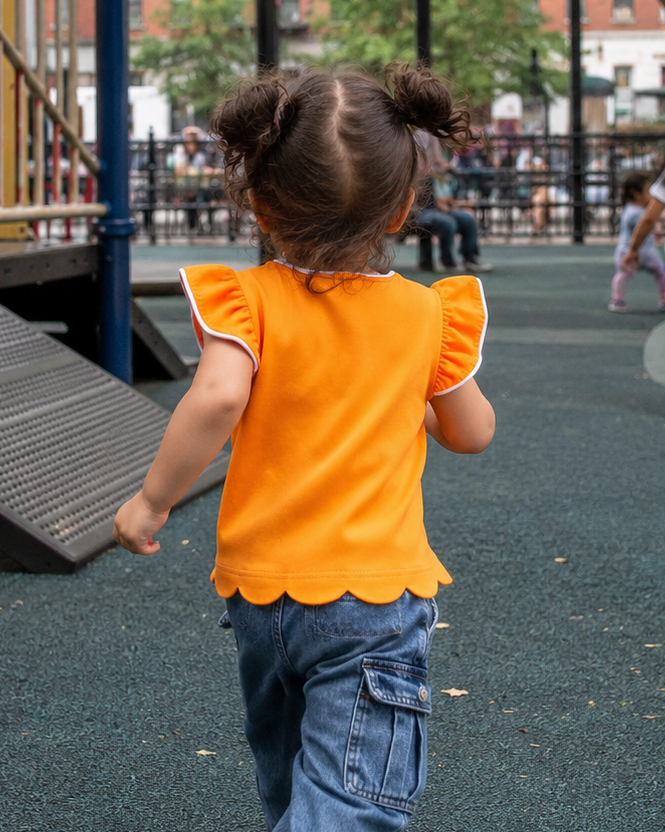 Orange kids flutter-sleeve top with white trim and scalloped hem, rear playground lifestyle view