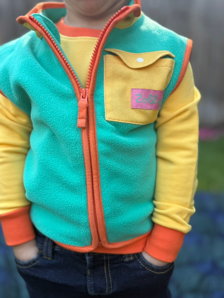 Teal toddler fleece vest with yellow chest pocket and orange zipper, modeled close-up front view